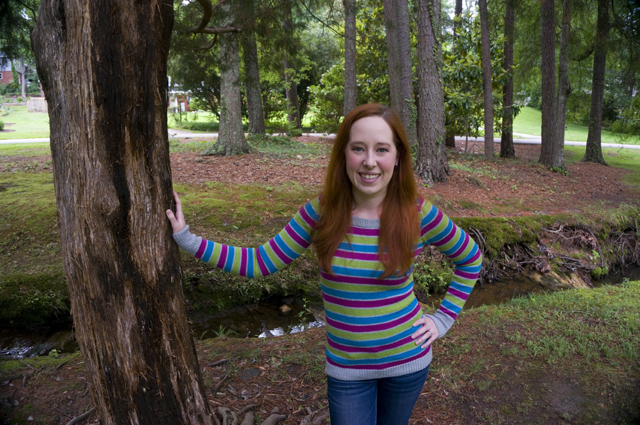 Caitlyn is standing in a local park with one hand on a tree and the other on her hip. She's wearing a striped sweater and jeans and smiling directly at the camera.