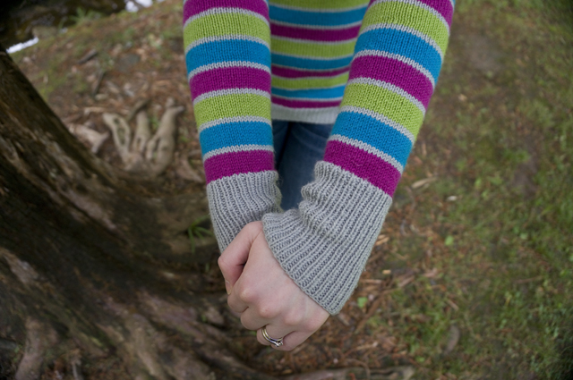 A close-up of Caitlyn's forearms and hands. Her striped sweater has wider fuchsia, turquoise, and lime stripes alternating with thinner light grey stripes, and light grey ribbed cuffs.