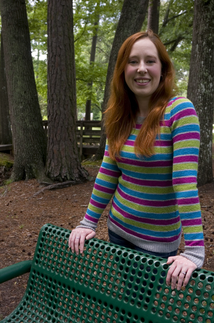 Caitlyn is resting her hands on the back of a green park bench and smiling at the camera. The stripes on her sweater aligned across the body and sleeves, and the ribbing on the cuffs is echoed at the hem.