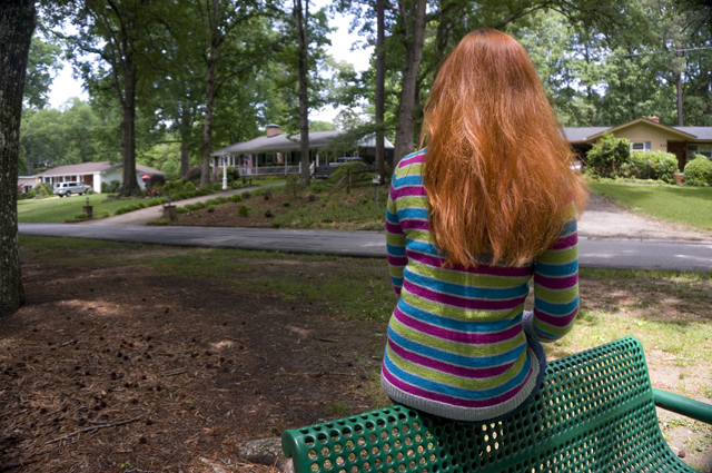 Caitlyn is sitting on a green park bench facing away from the camera toward houses in the distance.