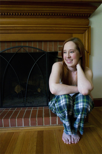 Caitlyn seated on a hearth, elbows resting on the knees of her tartan pajama pants, looking to the side and smiling