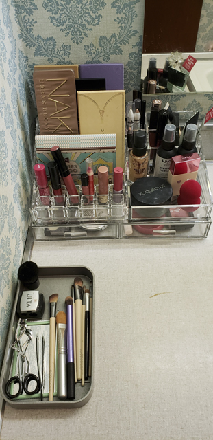 Stackable clear acrylic makeup storage holds a variety of lipsticks, eyeshadow palettes, and creams, and another plastic tray holds makeup brushes and cosmetic tools