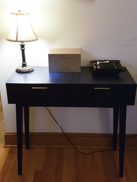 A shallow, two-drawer console table with brass pulls, topped by a short lamp, a gold lacquer box, and a leather-look key tray