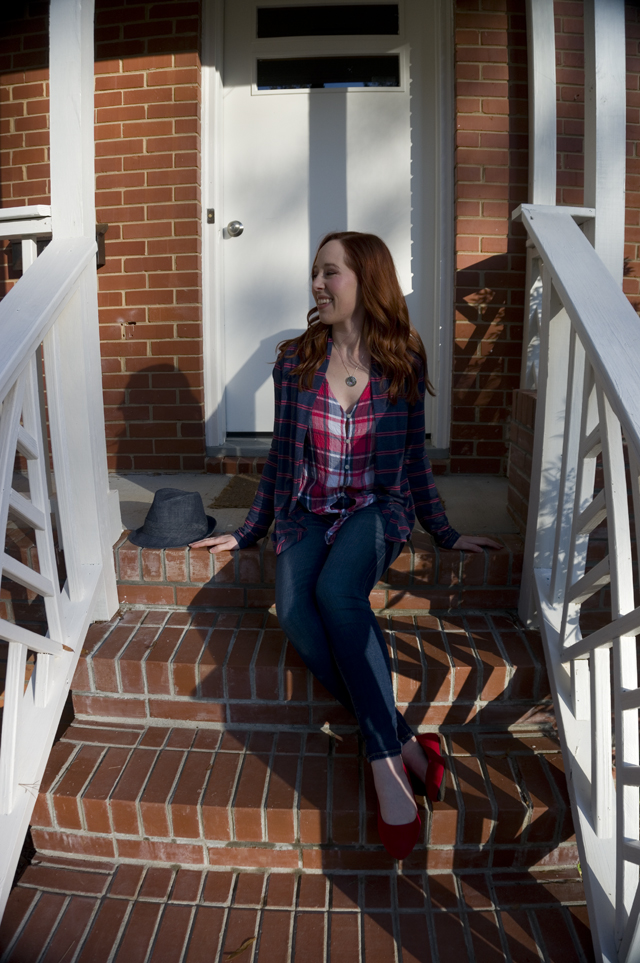 Caitlyn sits on her front porch next to a denim hat, smiling and wearing an open-front cardigan, tie-front button-up shirt, jeans, and pointed-toe flats