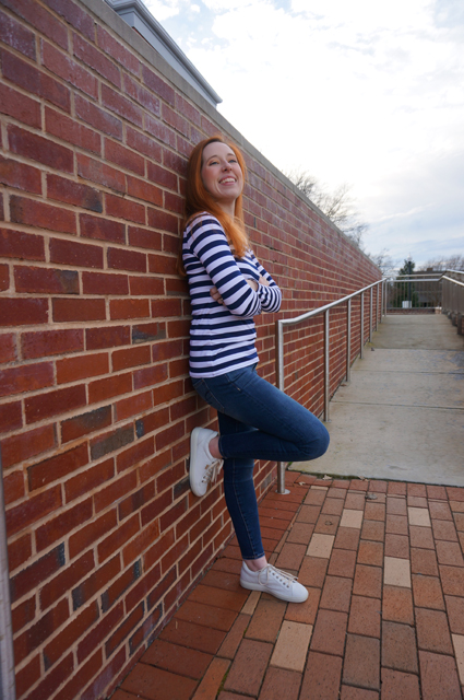 Caitlyn is standing with her back and one foot against a brick wall, arms crossed but smiling as she shows off the long-sleeve striped t-shirt she made