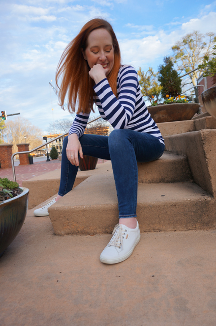 Caitlyn is wearing a long-sleeve striped t-shirt she made, sitting on the steps of a public building, and smiling with her eyes closed as though thinking of a secret