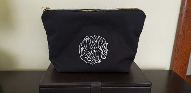 A black zippered pouch with outline of the words "Mind Fuzz" embroidered in white in a circle in the center of the pouch