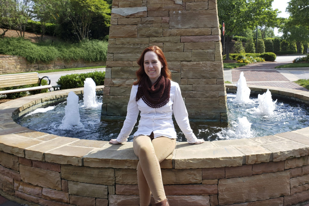 Caitlyn is sitting on a stone ledge around a fountain. She is wearing her Henslowe shawl wrapped around her neck like a bandana.